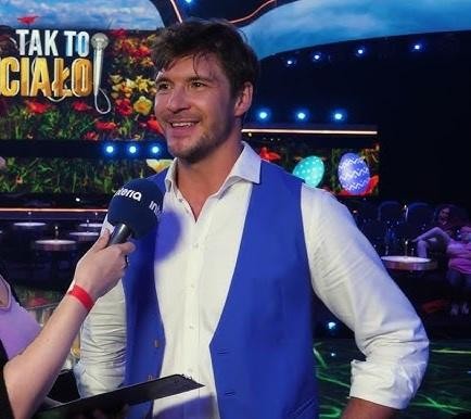 Mikołaj Roznerski na planie filmowym, wcielający się w rolę, z dynamicznym tłem sugerującym pracę aktorską.
