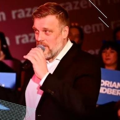 Adrian Zandberg przemawiający z pasją na wiecu politycznym, gestykulujący, z widocznym tłumem w tle.