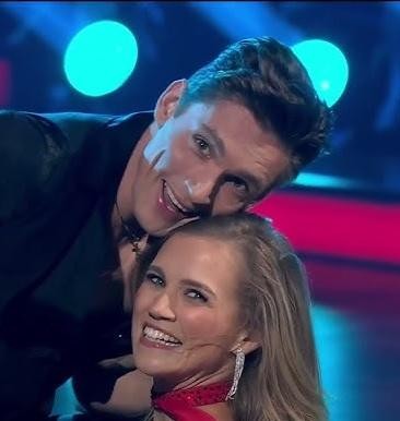 Agnieszka Kaczorowska w dynamicznej pozie tanecznej, w eleganckim kostiumie, na jasno oświetlonym parkiecie, z rozmytym tłem sugerującym ruch.