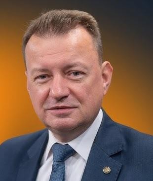 Mariusz Błaszczak przemawiający na tle polskiej flagi, symbolizujący jego rolę w polityce obronnej i krajowej.