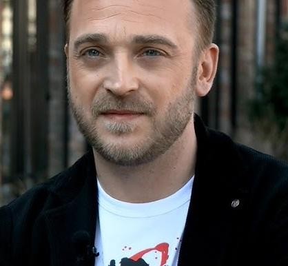 Mateusz Gessler w akcji w kuchni, ubrany w strój szefa kuchni, skupiony na gotowaniu lub prezentowaniu dania.