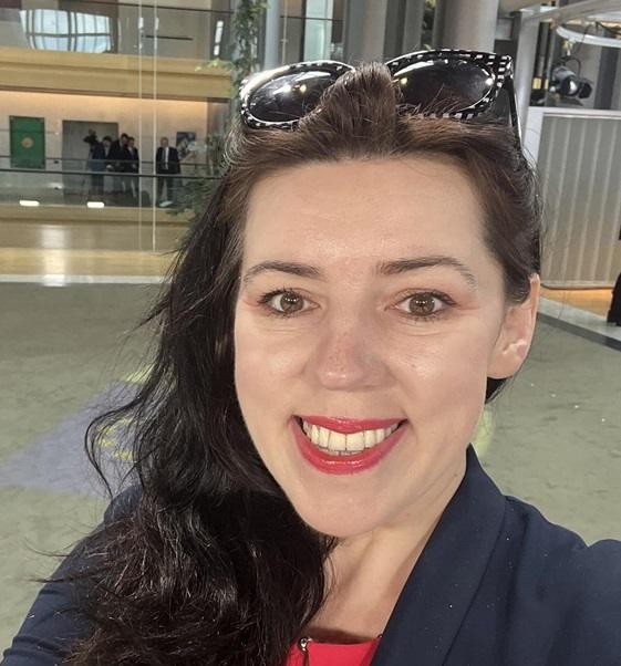 Dominika Ćosić w roli dziennikarki, z mikrofonem lub notatnikiem, w profesjonalnym otoczeniu, symbolizującym jej pracę w polityce międzynarodowej.