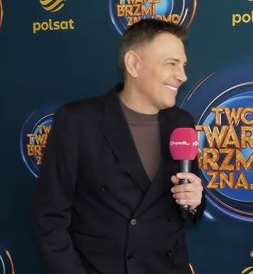 Krzysztof Ibisz w studiu telewizyjnym, uśmiechnięty i pełen energii, w tle widoczne elementy scenografii telewizyjnej.